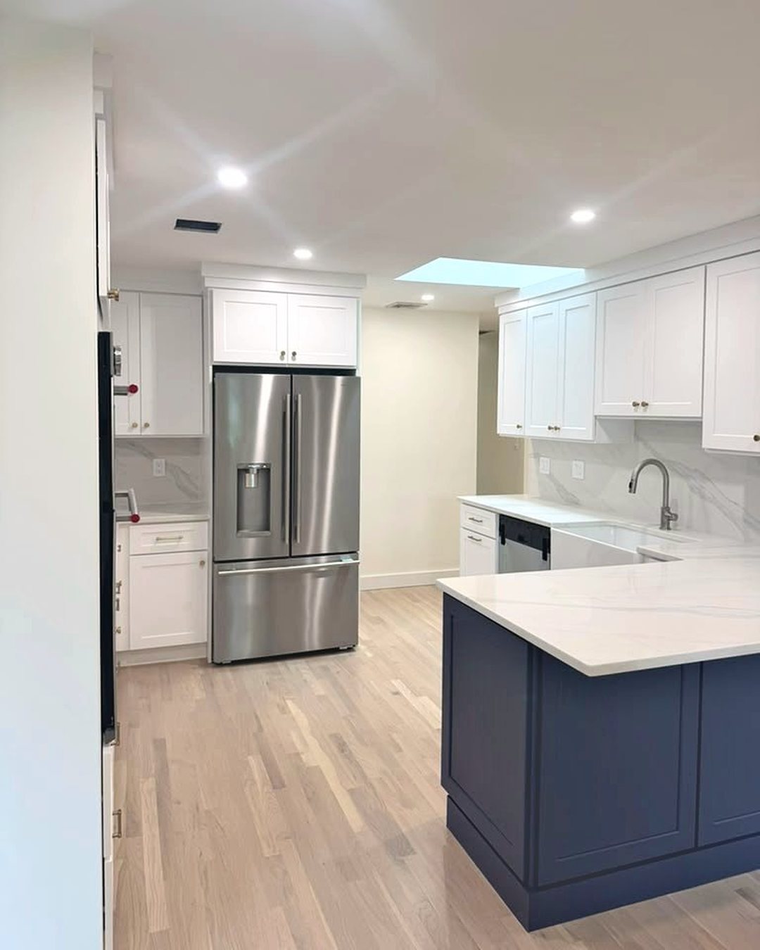 The same kitchen finished: white shaker cabinets, navy peninsula, quartz counters, refinished oak floor