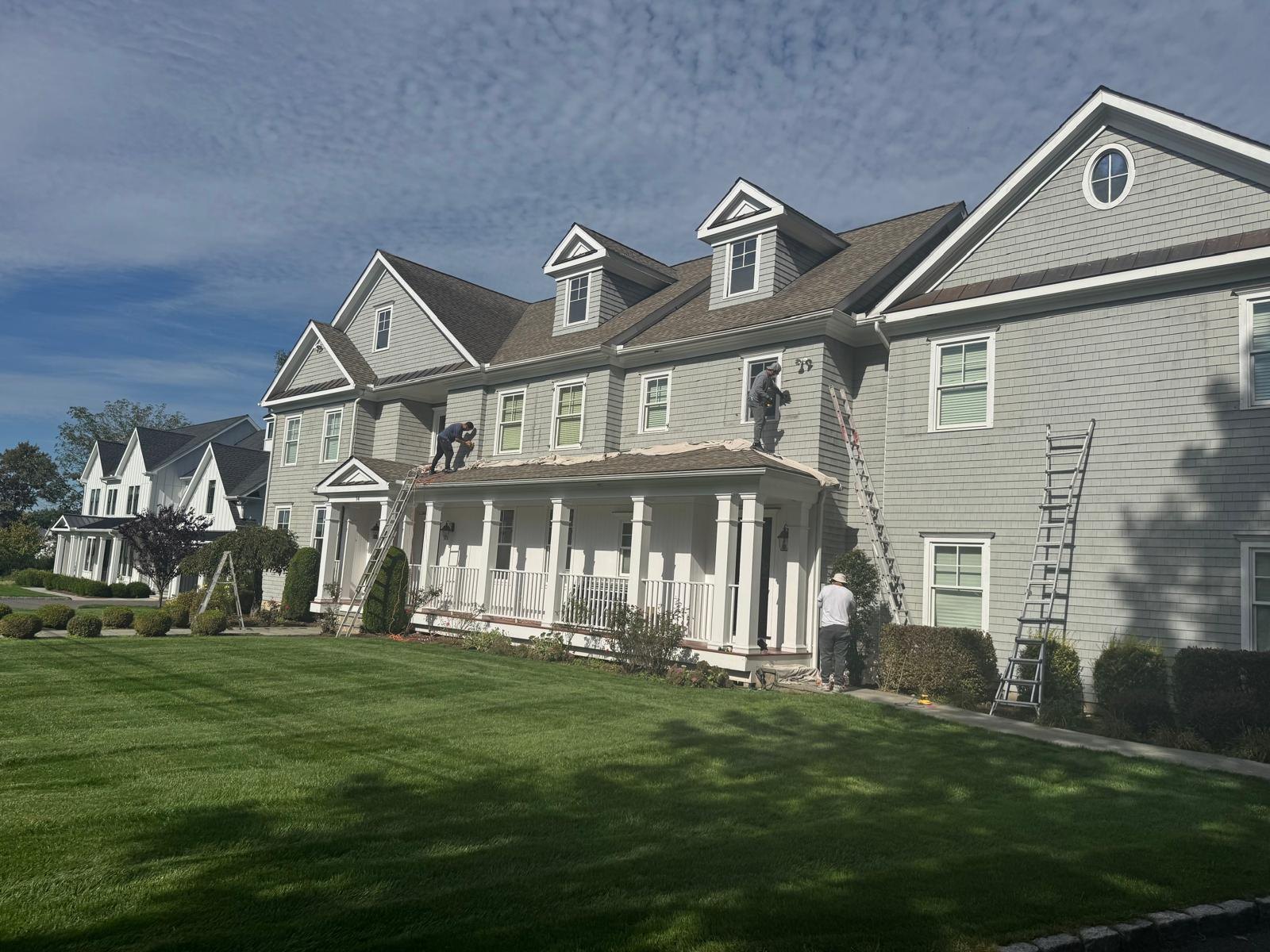 Gray shingle house with a white porch, crew on the porch roof and ladders before painting